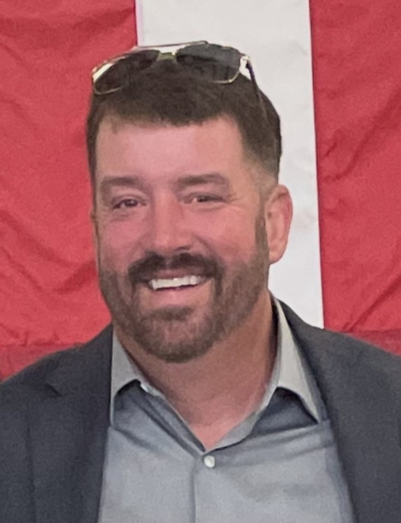 A smiling man with short dark hair and a beard, wearing sunglasses on his head and a button-up shirt, in front of a red and white background.